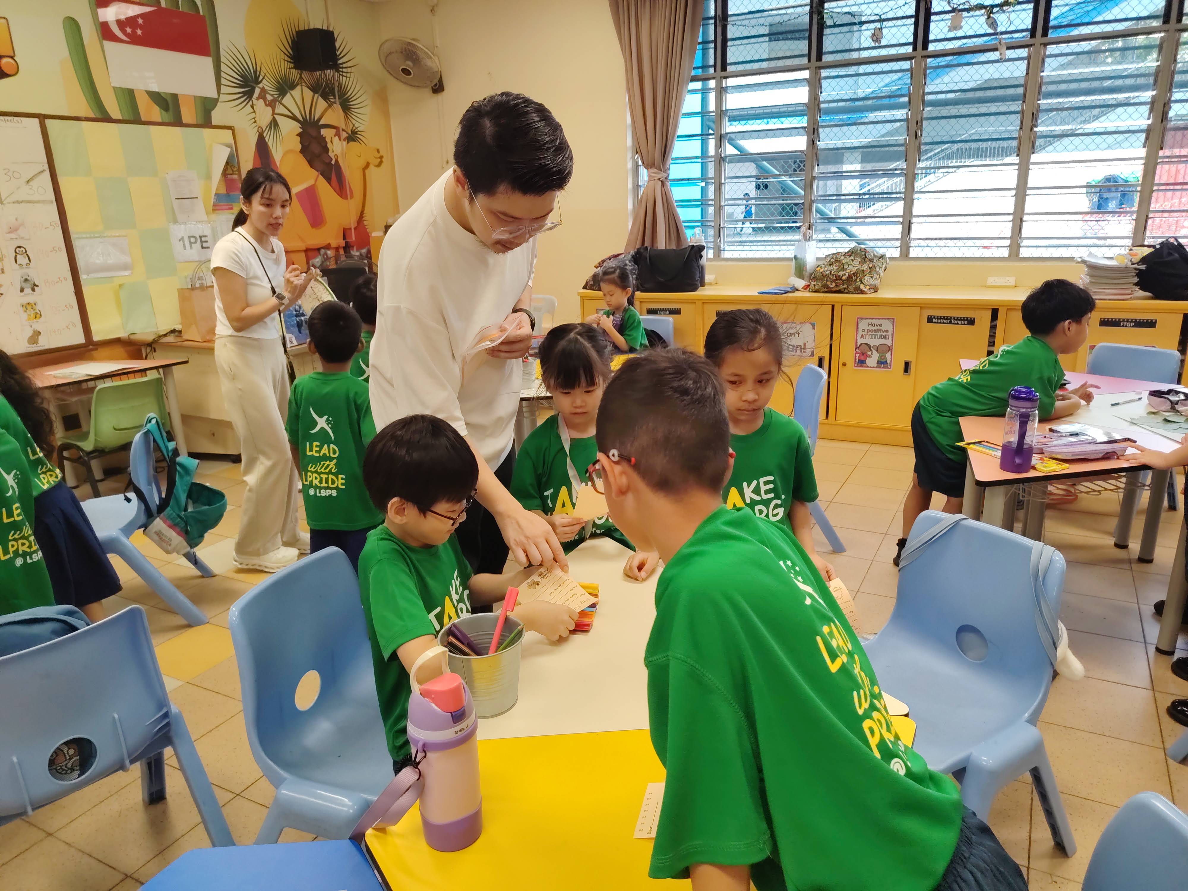 A parent volunteer leans over a table to assist a group of young students in green school T-shirts during a classroom activity. Another parent volunteer stands in the background as students work on their tasks around the classroom.